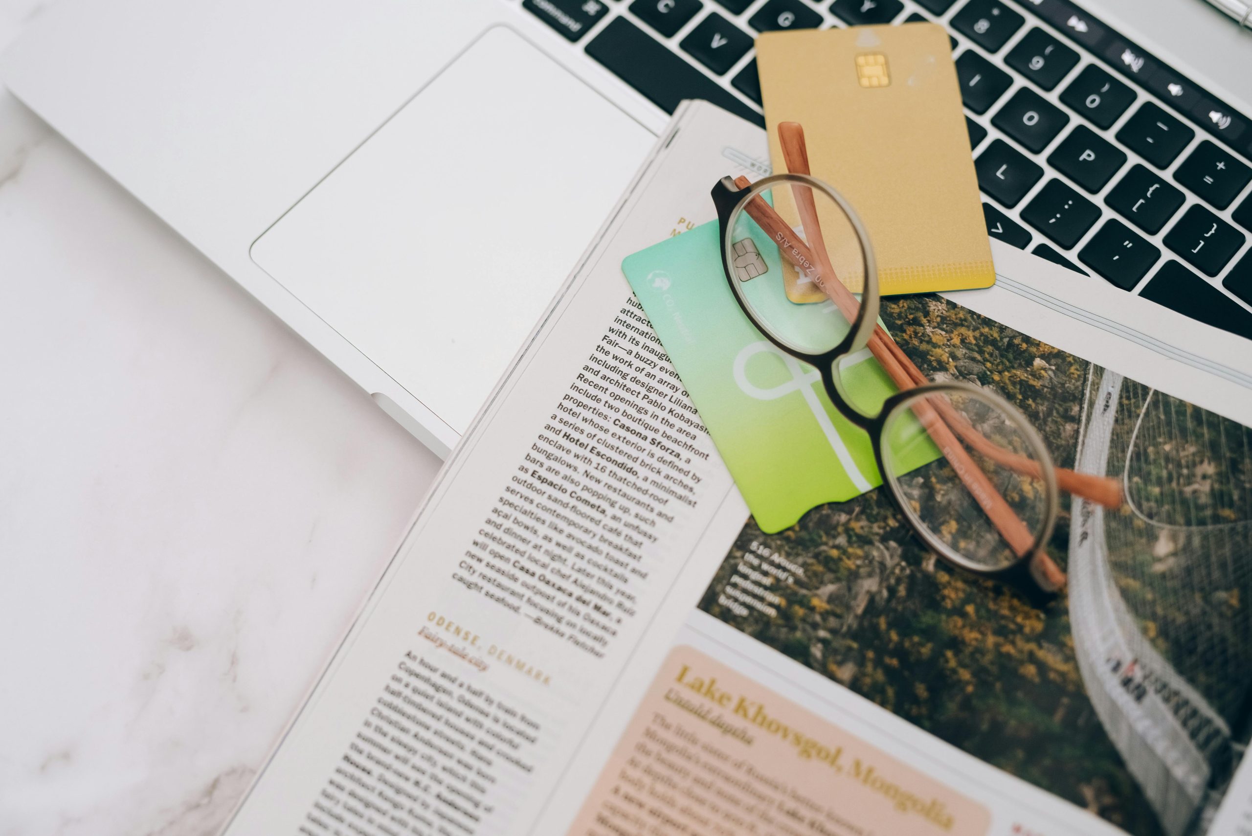 Eyeglasses resting on a magazine beside a laptop and credit cards, indoors.
